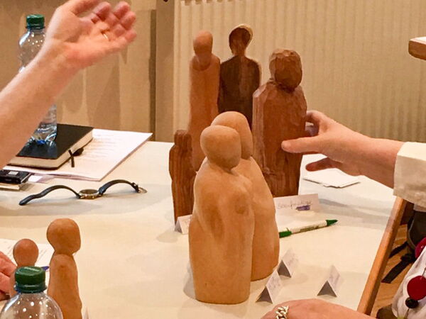 Sie sehen verschiedene Holzfiguren auf einem Tisch, mit Beschriftungsschildchen. Es wird gerade damit gearbeitet, was man an Händen sieht, die diese gerade anfassen und bewegen.